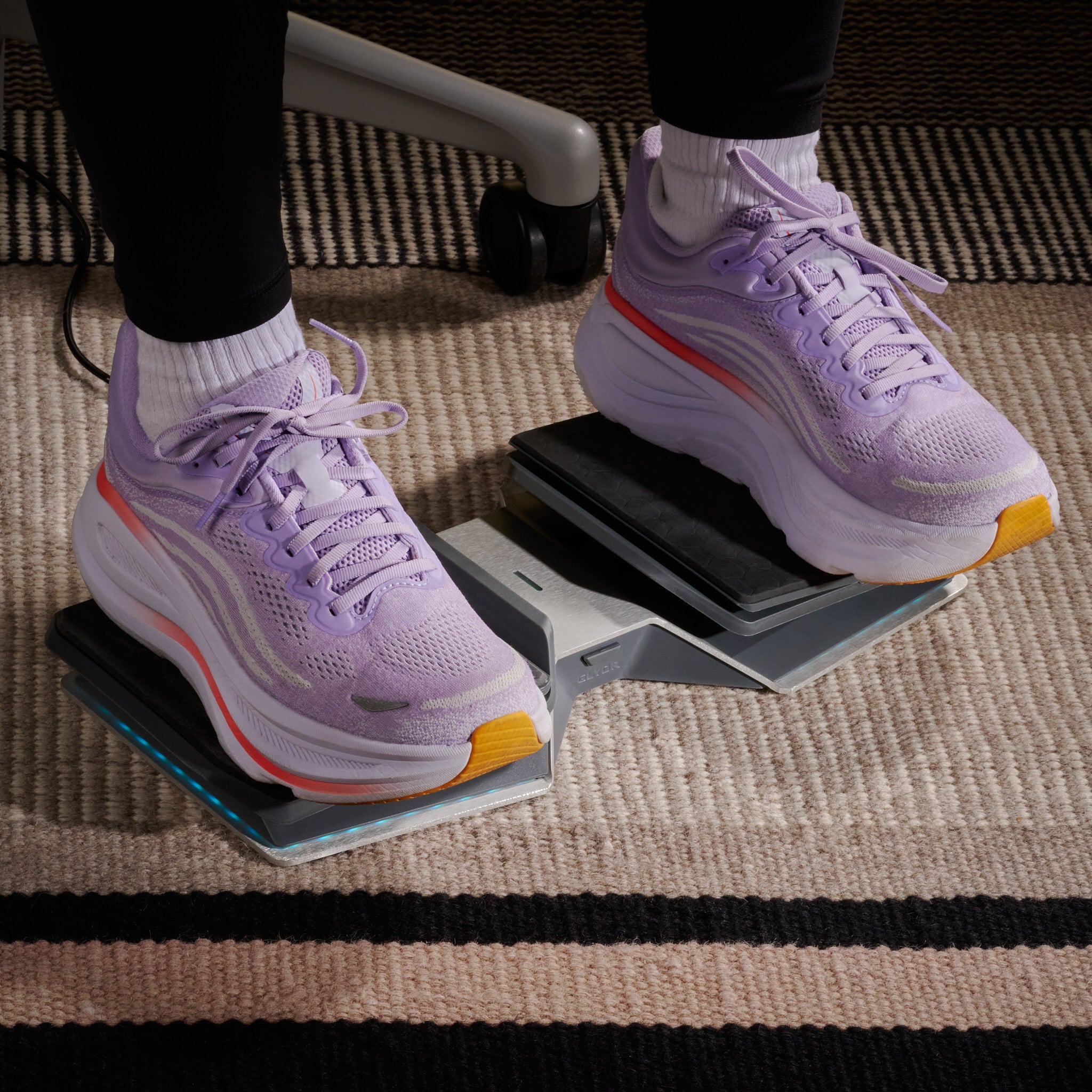 A person’s feet in light purple sneakers placed flat on a each pedal with a striped rug in the background